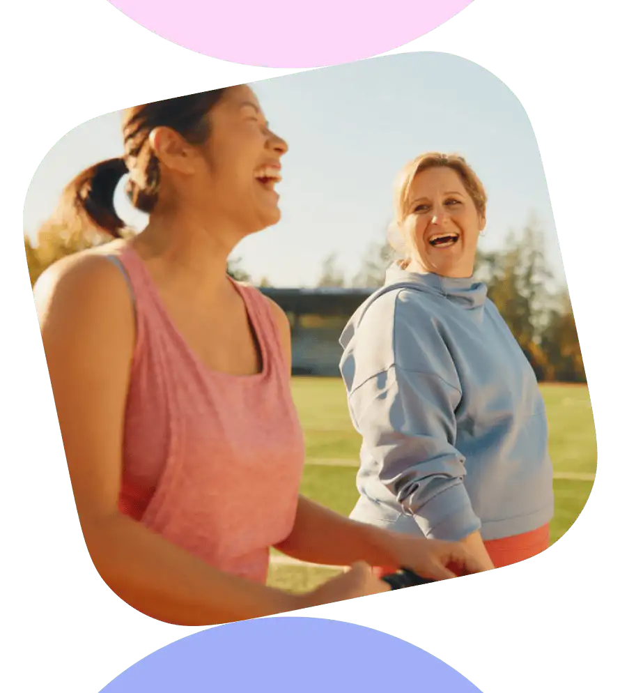 Two women laughing and smiling in a sunlit field, enjoying each other's company on a beautiful day.